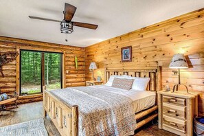 A log cabin bedroom with a bed, bedside tables, a ceiling fan, and a large window. - Lakefront log cabin on 3.5 acres with dock, gazebo & sunset views - dog-friendly (Lac Du Flambeau)