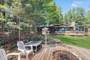 A wooden deck with a table and chairs, a stone fire pit, and a cabin with a deck and glass windows.