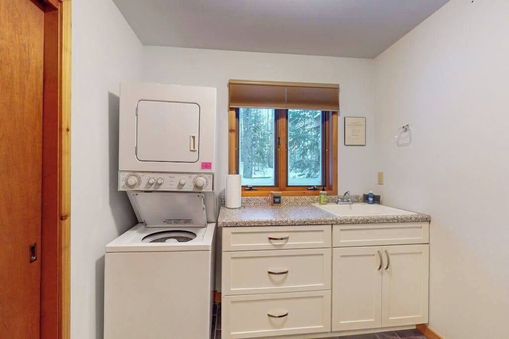 Een compacte keuken met een ingebouwde wasmachine en droger, een raam met uitzicht op bomen, en een wastafel met aanrecht.