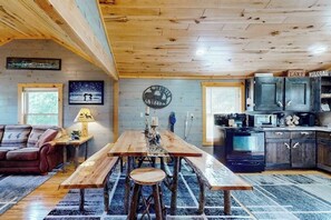 A rustic cabin interior with a wooden dining table, a sofa, a kitchen area, and a wall-mounted clock. - Cozy home located near Lakes & Trails equipped with WIFI & a Gas Grill (Mercer)