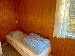 Ein Einzelbett mit gesteppter Matratze und Kissen, vor einer Holzwand mit einem Fenster und weißem Vorhang.