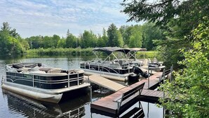 A docked pontoon boat with a covered seating area and a wooden dock extending into a calm lake surrounded by dense forest. - Cozy Cabin located on the Turtle Flambeau Flowage, great for all seasons! (Mercer)