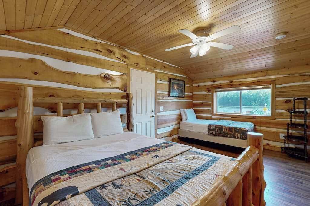 A log cabin bedroom with a bed, a bench, a ceiling fan, and a window.