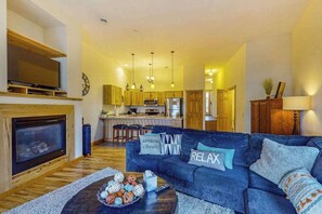 A living room with a fireplace, a sofa with decorative pillows, a coffee table with a bowl of fruit, and a kitchen area in the background.