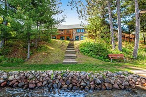 A building with a stone pathway leading to a wooden deck, surrounded by trees and a stone wall.