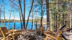 En campingplats med en eldstad, trästolar och utsikt över en sjö som omges av träd.