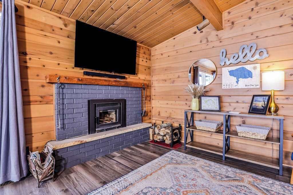 Interior de una cabaña de troncos con chimenea, televisor montado, un letrero que dice 'hello' y un mapa enmarcado en la pared.