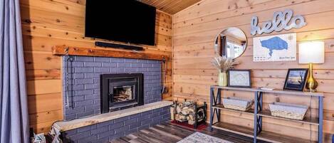 Interior de una cabaña de troncos con chimenea, televisor montado, un letrero que dice 'hello' y un mapa enmarcado en la pared.