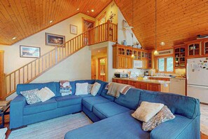 A modern living room with a blue sectional sofa, wooden staircase, and a kitchen area. - Serene Lakefront Home w/ Wifi, dock, kayaks, gas grill, fire pit & more (Eagle River)