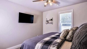 Ein Schlafzimmer mit einem Bett, einem Deckenventilator, einem an der Wand befestigten Fernseher, einem Fenster mit Jalousien und einem Wandregal mit Dekorationen.