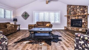 Ein Wohnzimmer mit Kamin aus Stein, Deckenventilator und großem Fenster mit Jalousien.