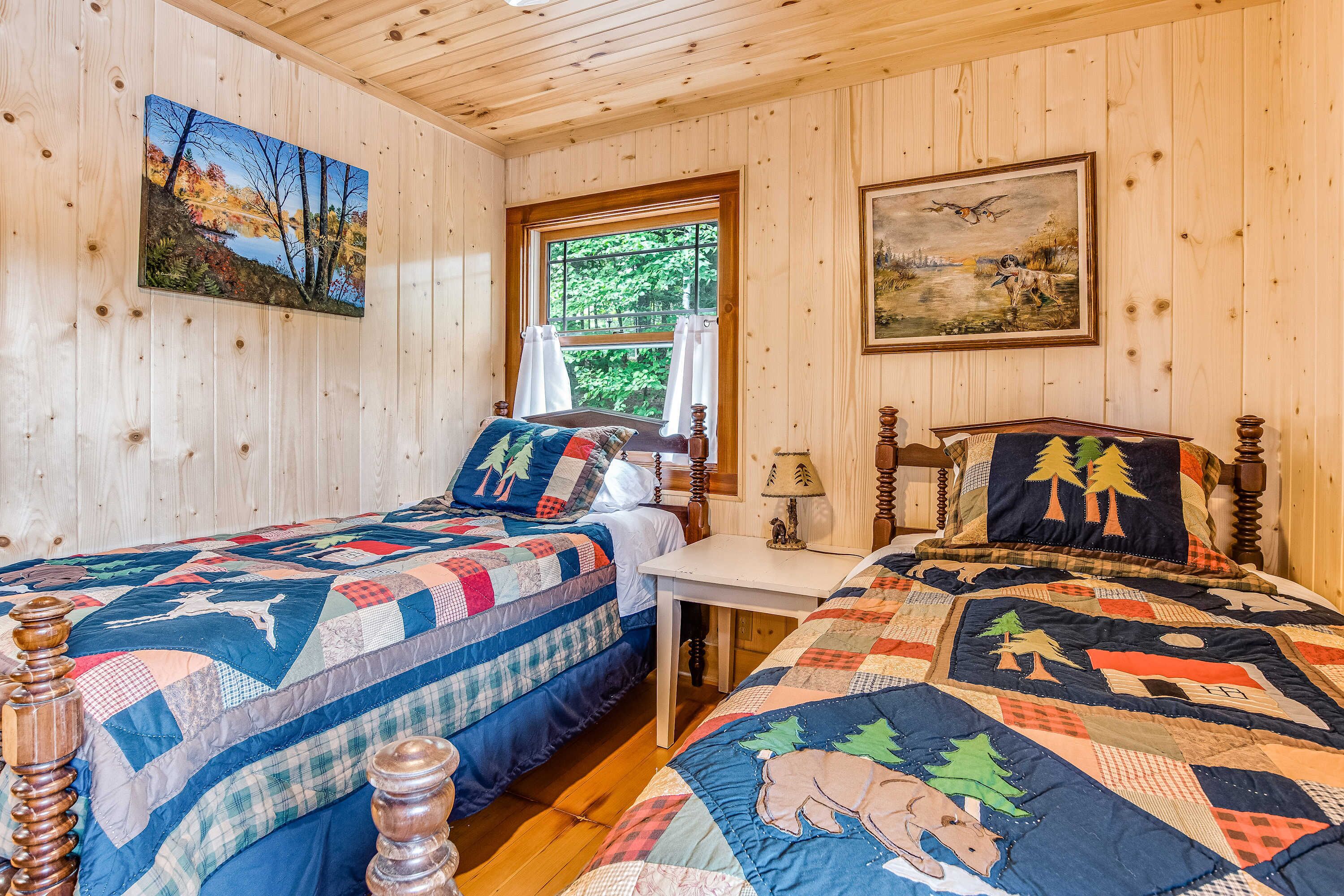 A cabin-style bedroom with two wooden beds, a window with curtains, and two paintings on the walls.