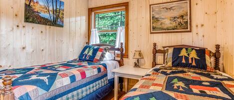 A cabin-style bedroom with two wooden beds, a window with curtains, and two paintings on the walls.