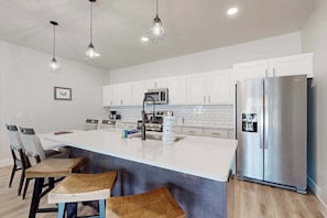 Cocina moderna con armarios blancos, refrigerador de acero inoxidable, una isla central con taburetes y iluminación colgante.