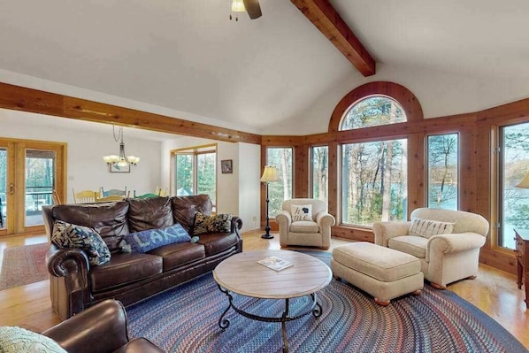 A spacious living room with a brown leather sofa, two armchairs, a round coffee table, and large windows offering a view of trees.