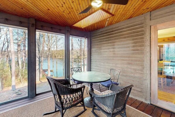 A covered patio with a round table and wicker chairs, large windows, and a wooden ceiling.