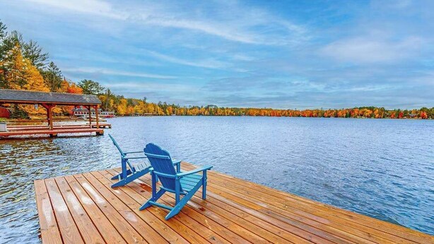 Eine Holzsteg mit zwei blauen Adirondack-Stühlen, die auf einen stillen See ausgerichtet sind, umgeben von Herbstlaub.