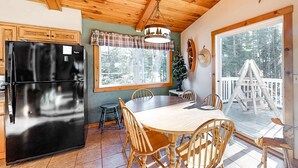 Eine Küche mit einem schwarzen Kühlschrank, einem Holzespeisesaal mit Stühlen, einem Fenster mit Karomuster-Vorhang und einer Terrasse mit Holzwertlauf.