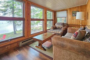 A cozy cabin interior with large windows, a wooden table, and a sofa with patterned cushions. - Waterfront home between 2 lakes w/ heated porch, dock & kayaks (Manitowish Waters)