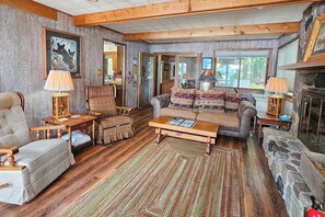A cozy living room with wooden floors, a fireplace, and a large window. - Waterfront home between 2 lakes w/ heated porch, dock & kayaks (Manitowish Waters)