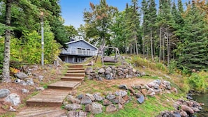 Eine Holzhütte mit einer Veranda und einer Holzterrasse, umgeben von Wald und einem Bach.