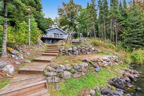 A cabin with a wooden deck leading to a porch, surrounded by a forest and a stream. - Beautiful home on High Lake w/private pier, large deck & canoes (Land O'Lakes)