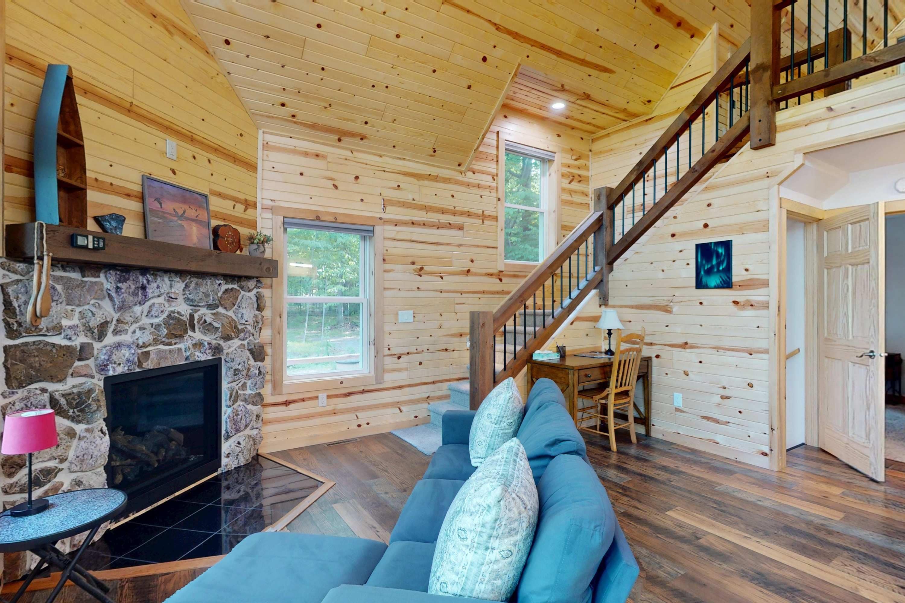 A cozy cabin interior with a stone fireplace, a blue sofa, a wooden staircase, and a dining area.