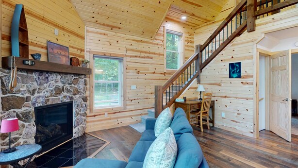 A cozy cabin interior with a stone fireplace, a blue sofa, a wooden staircase, and a dining area. - Lakefront Sunrise Lodge: fireplace, deck views, kayaks, pier & firepit (Rhinelander)