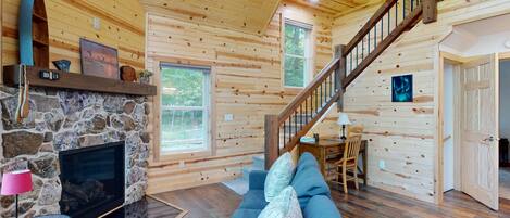 Interior acogedor de una cabaña con chimenea de piedra, un sofá azul, una escalera de madera y un comedor.