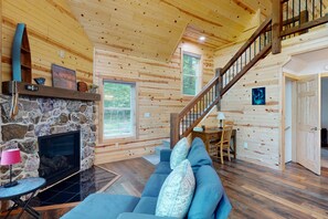 A cozy cabin interior with a stone fireplace, a blue sofa, a wooden staircase, and a dining area.