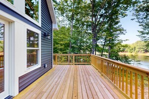 A wooden deck with a railing, a window, and a view of a lake surrounded by trees.