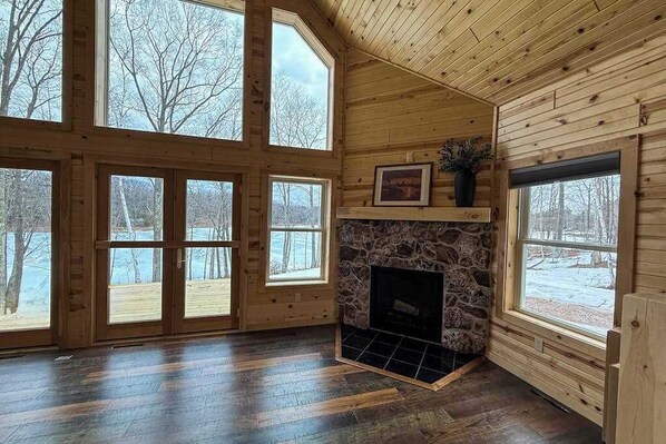 A log cabin interior with a stone fireplace, large windows, and a view of the outdoors.