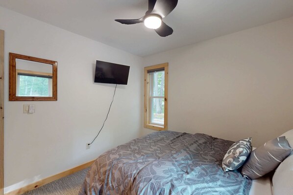 A bedroom with a bed, a ceiling fan, a TV mounted on the wall, and two windows.