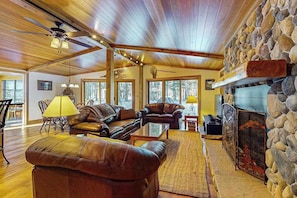 A spacious living area with leather sofas, a stone fireplace, and large windows. - Charming Waterfront Home w/ Dock, Fire pit, WIFI, near Town & Trails (Eagle River)