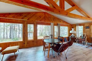 Une cabane avec un plafond haut, des poutres en bois, de grandes fenêtres, une table à manger, des chaises, un canapé et une radio.