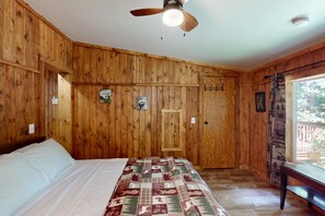 A room with wooden paneling, a bed with a quilt, a ceiling fan, and a window with a view of trees.