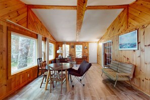 A wooden interior with a dining table, chairs, a bench, and large windows.