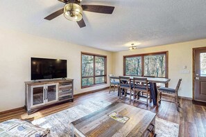 A spacious living room with a large TV, wooden furniture, and large windows offering a view of trees.