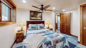 A bedroom with a bed, a ceiling fan, a nightstand with a lamp, a painting on the wall, and a door. - Cozy Wooded home with WIFI, Gas Grill, Two Kitchens, Near Lakes & Trails (Eagle River)