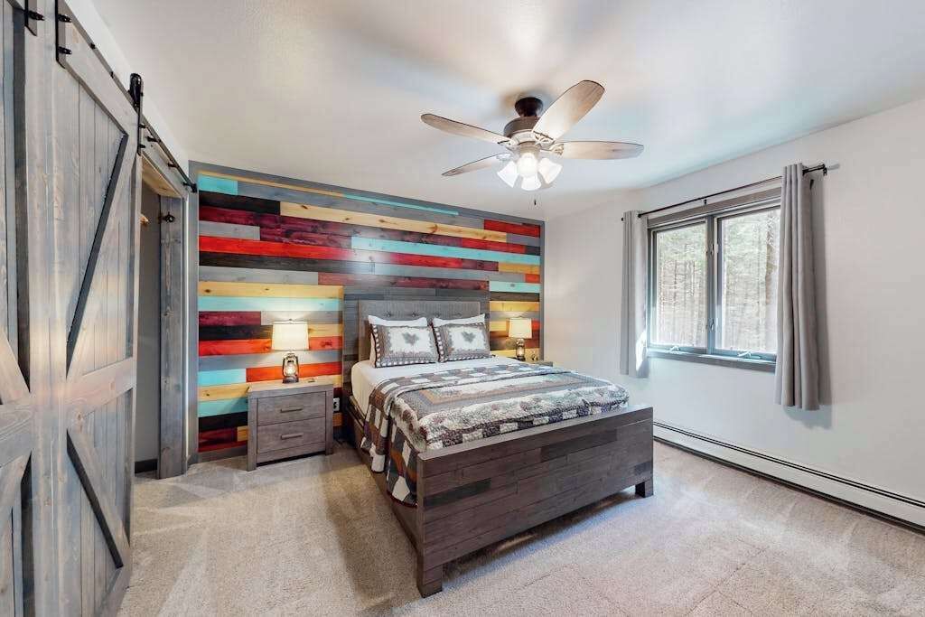A bedroom with a bed, a nightstand, a ceiling fan, a window, and a wall with a colorful panel design.