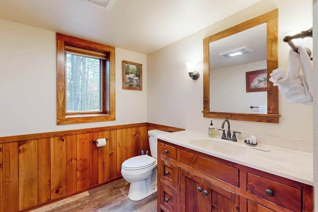 A bathroom with wooden paneling, a white toilet, a vanity with a mirror, and a window with a view of trees.