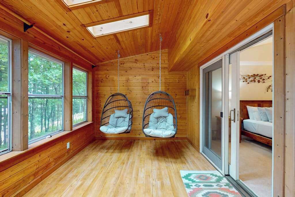Une pièce aux murs en bois, avec de grandes fenêtres, un lit et deux fauteuils suspendus.