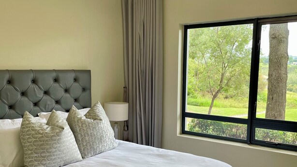 Une chambre à coucher avec une tête de lit capitonnée, un grand lit avec une literie blanche, une table de chevet avec une lampe et une fenêtre donnant sur un paysage verdoyant.