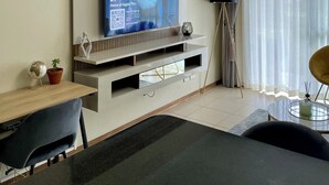 Un salon moderne avec un téléviseur à écran plat fixé au mur, un coin repas comprenant une table et des chaises, et une grande fenêtre ornée de rideaux légers.