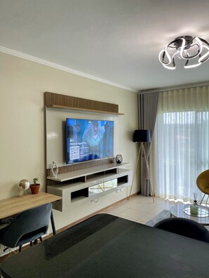 Ein modernes Wohnzimmer mit einem an der Wand befestigten Flachbildfernseher, einem Essbereich mit Tisch und Stühlen sowie einem großen Fenster mit durchscheinenden Vorhängen.