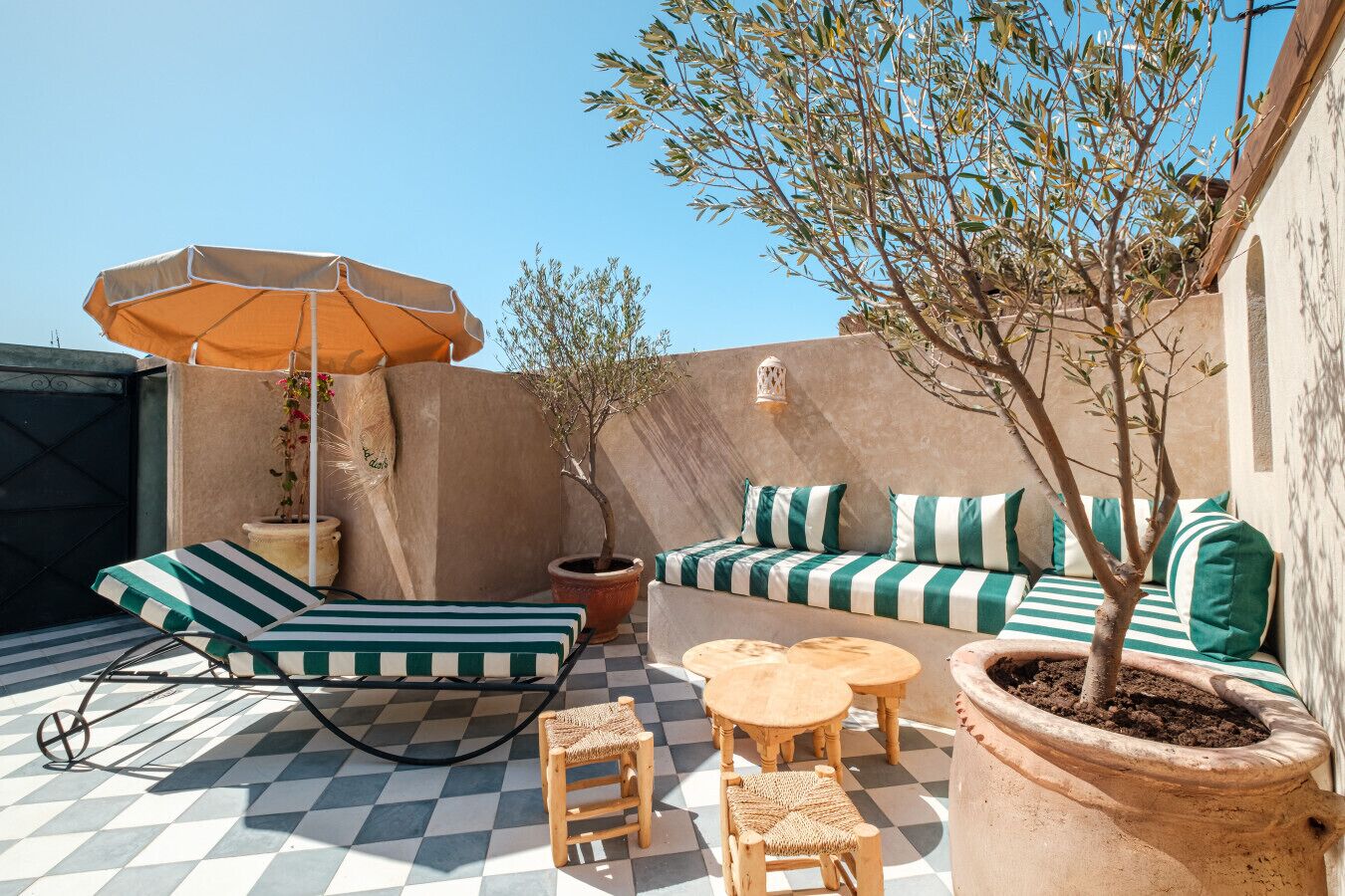 A rooftop terrace with a checkered floor, a striped sofa set, a wooden table, a potted tree, and an umbrella.