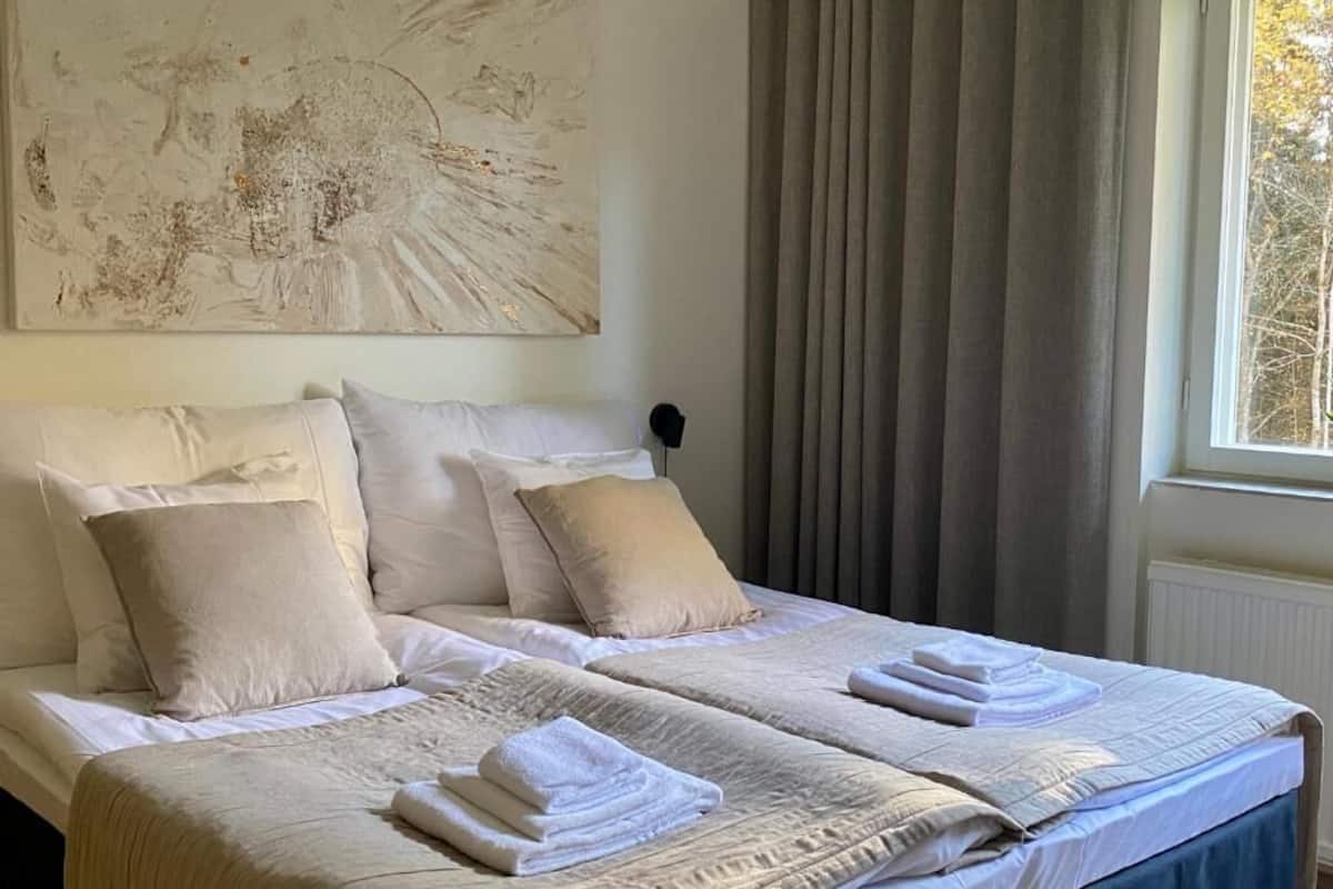 Una cama doble con ropa de cama color beige, un cuadro abstracto grande en la pared y una ventana con cortinas.