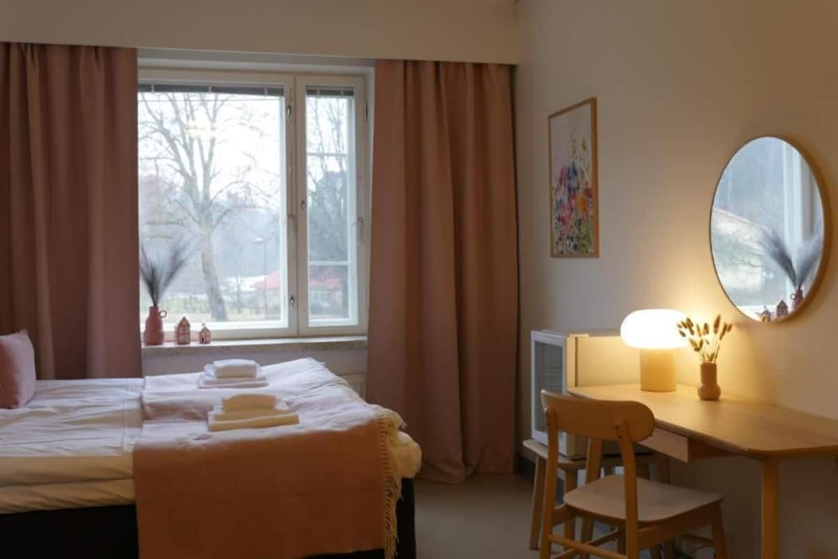 Habitación de hotel con cama, escritorio con lámpara, silla, televisor y ventana con cortinas.