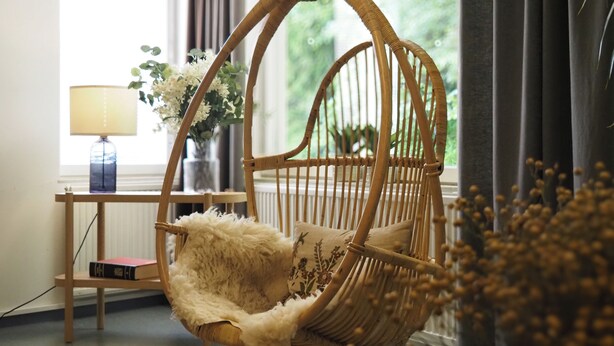 A wicker hanging chair with a fluffy cushion in a room with a window, a lamp, and a small table with a book and flowers. - EnjoyNature Resort (Hollola)