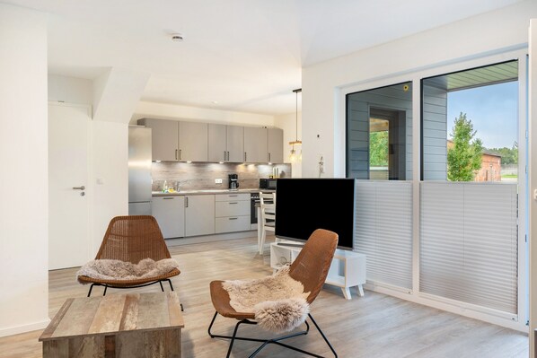 A modern kitchen with white cabinets, a dining area with two wicker chairs and a wooden table, and a large TV on a stand.